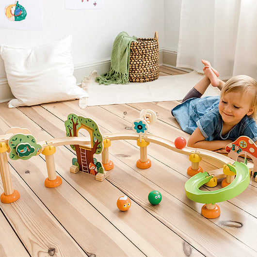 A child playing with colorful wooden pipe building blocks on a wooden floor. The blocks are shaped as animals and objects, and there is a small wooden structure with a ramp and decorative elements.