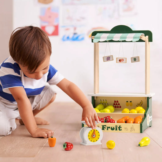 Child playing with a toy fruit stand and fruits on a beige floor.