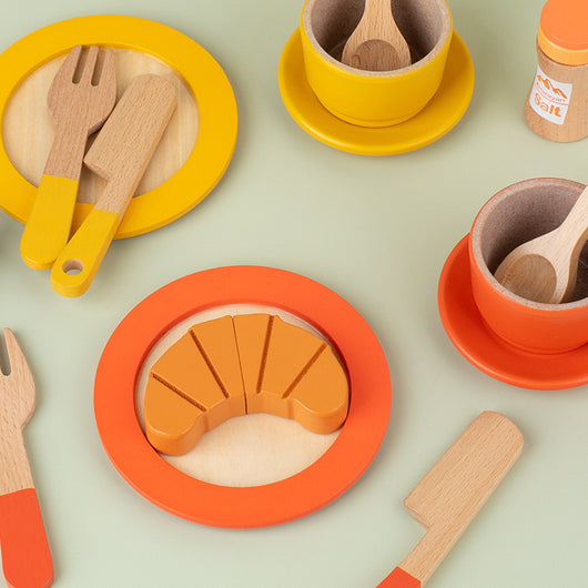 Colorful toy kitchen set with plates, cups, utensils, and a bread shape on a light green background.