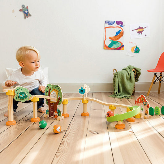 A young child playing with colorful wooden pipe building blocks on a wooden floor, with toys such as balls and a car nearby.