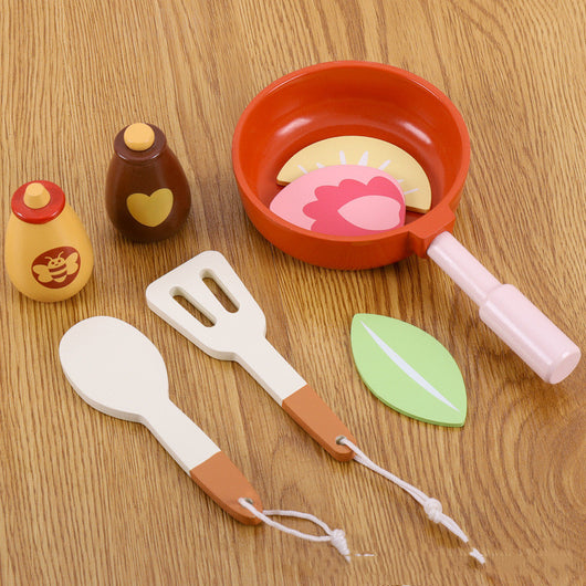 Set of toy kitchen utensils including a frying pan, spatula, and bottles on a wooden surface.