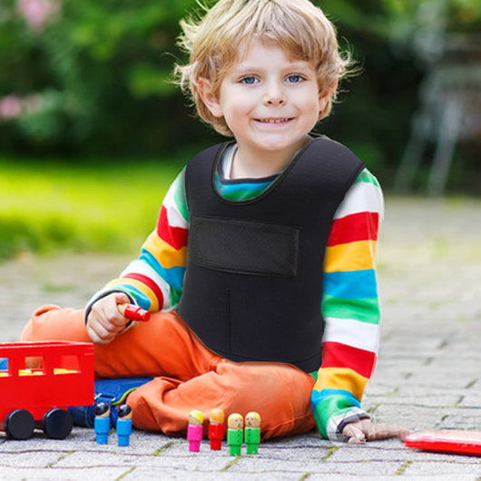 A child sitting on the ground wearing a black compression vest with adjustable straps, colorful striped shirt, and orange pants, playing with toy blocks and a toy train.