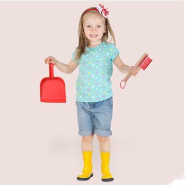 Child wearing a light blue shirt with colorful polka dots, denim shorts, yellow rain boots, and holding a red shovel and brush on a pink background.